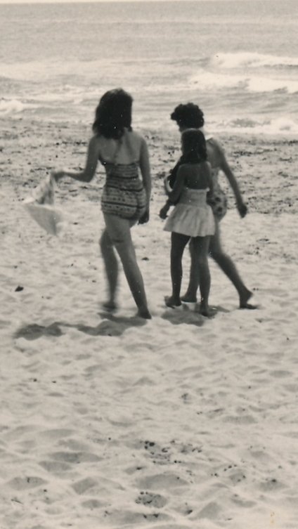 Three women walking on a sandy beach, two wearing swimsuits and one in a skirt, enjoying a sunny day by the ocean.